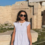 Woman in a white dress and sunglasses stands in front of ancient stone ruins on a sunny day. She is smiling slightly, with her hair down, and there is greenery and blue sky in the background.