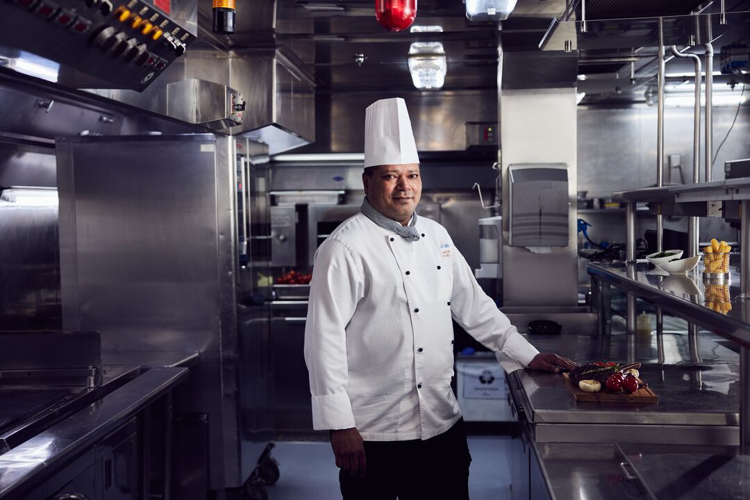 A chef in a white uniform and tall hat stands in a modern, stainless steel kitchen, resting one hand on a worktop where a wooden board holds plated food, including tomatoes and greens.