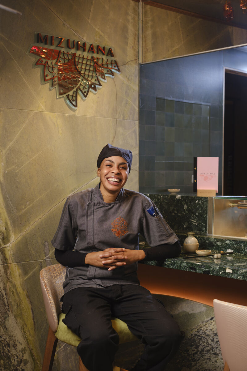 A smiling chef in a dark uniform and hat sits on a chair at a counter in a modern restaurant with a sign that reads Mizuhana on the wall behind them.
