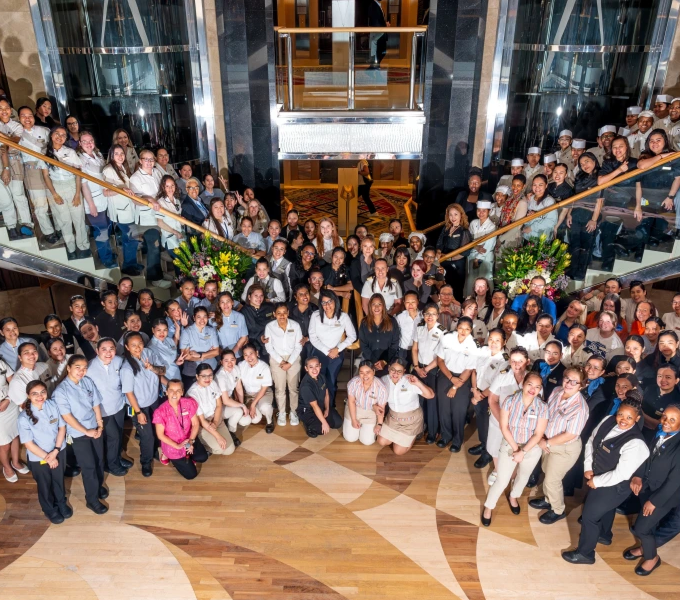 A large, diverse group of uniformed staff members poses together in a spacious, elegant lobby with stairs, flowers, and a lift in the background, smiling for a group photo.