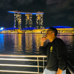 A person stands by a railing at night overlooking a lit waterfront with the Marina Bay Sands hotel illuminated in the background in Singapore.