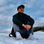 A person wearing a dark jacket, grey trousers, white trainers, and a black cap sits on snowy ground with a cloudy blue sky in the background.