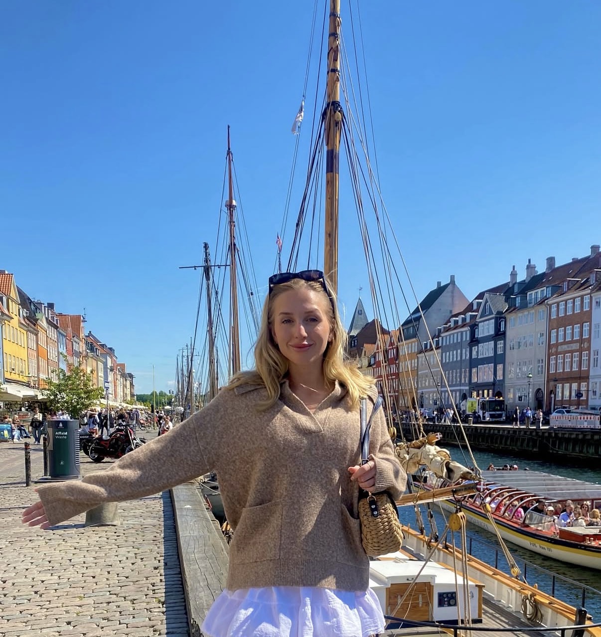 A woman in a beige jumper and white skirt smiles with one arm outstretched by a waterfront, colourful buildings and moored sailboats behind her—evoking the adventure of nursing careers at sea under a clear blue sky on a sunny day.