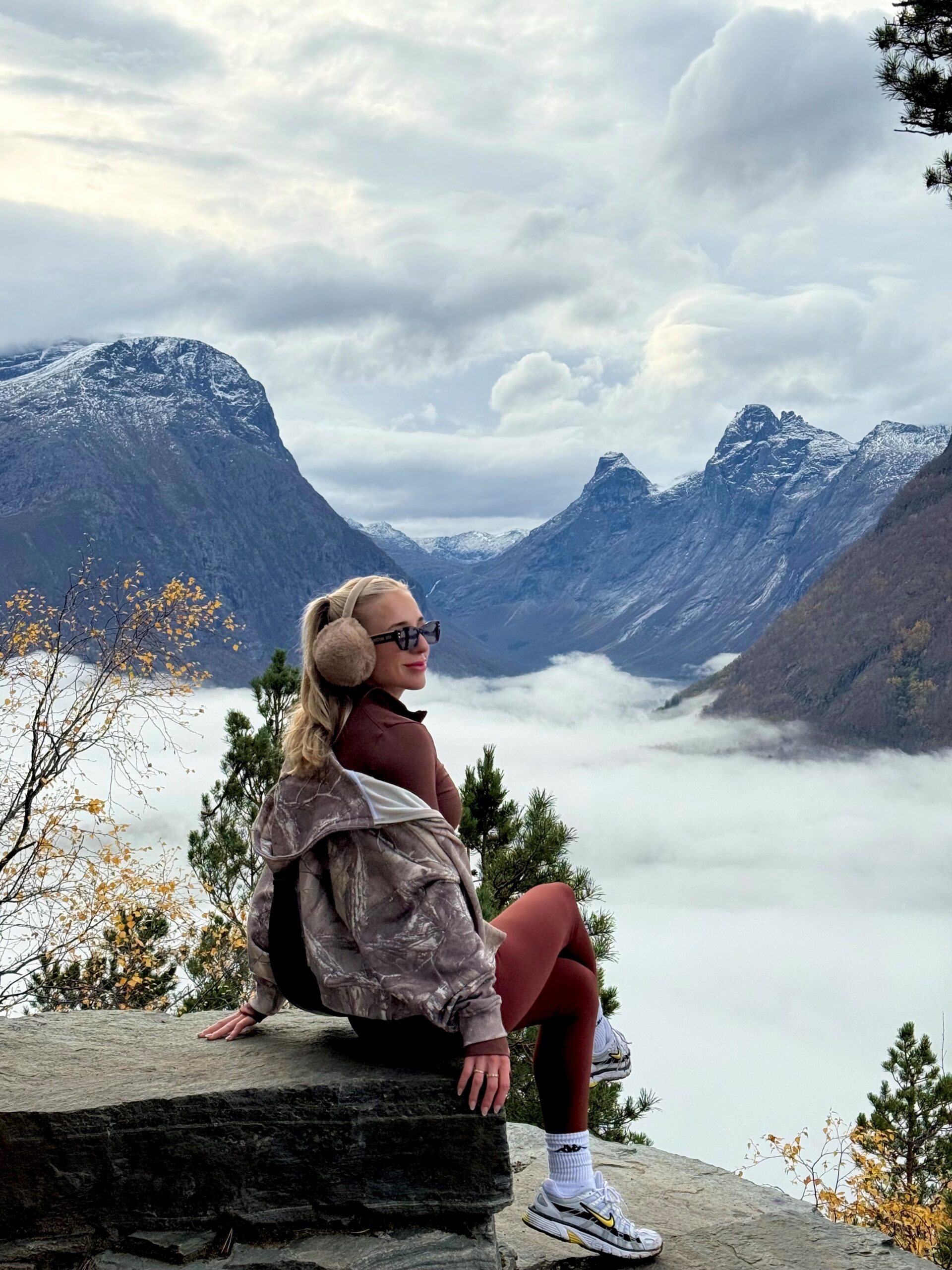 A woman in earmuffs, sunglasses, and sportswear sits on a stone ledge, smiling, with misty mountains behind her—much like those exploring nursing careers at sea enjoy breathtaking views on their journeys.