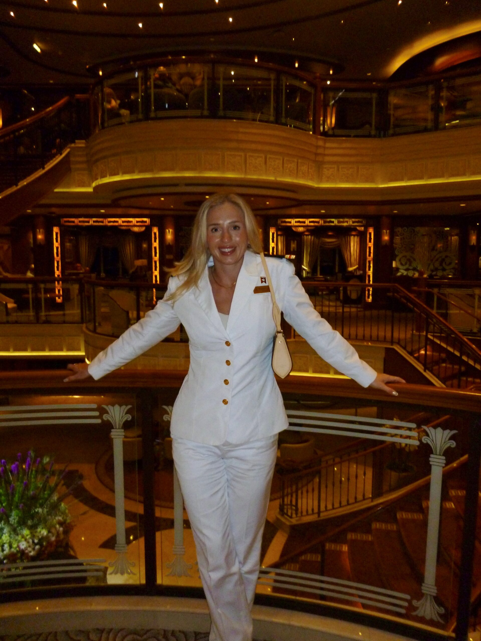 A woman with long blonde hair, dressed in a white suit, stands smiling with arms resting on a decorative railing—her elegance reflecting the rewarding opportunities found in nursing careers at sea.