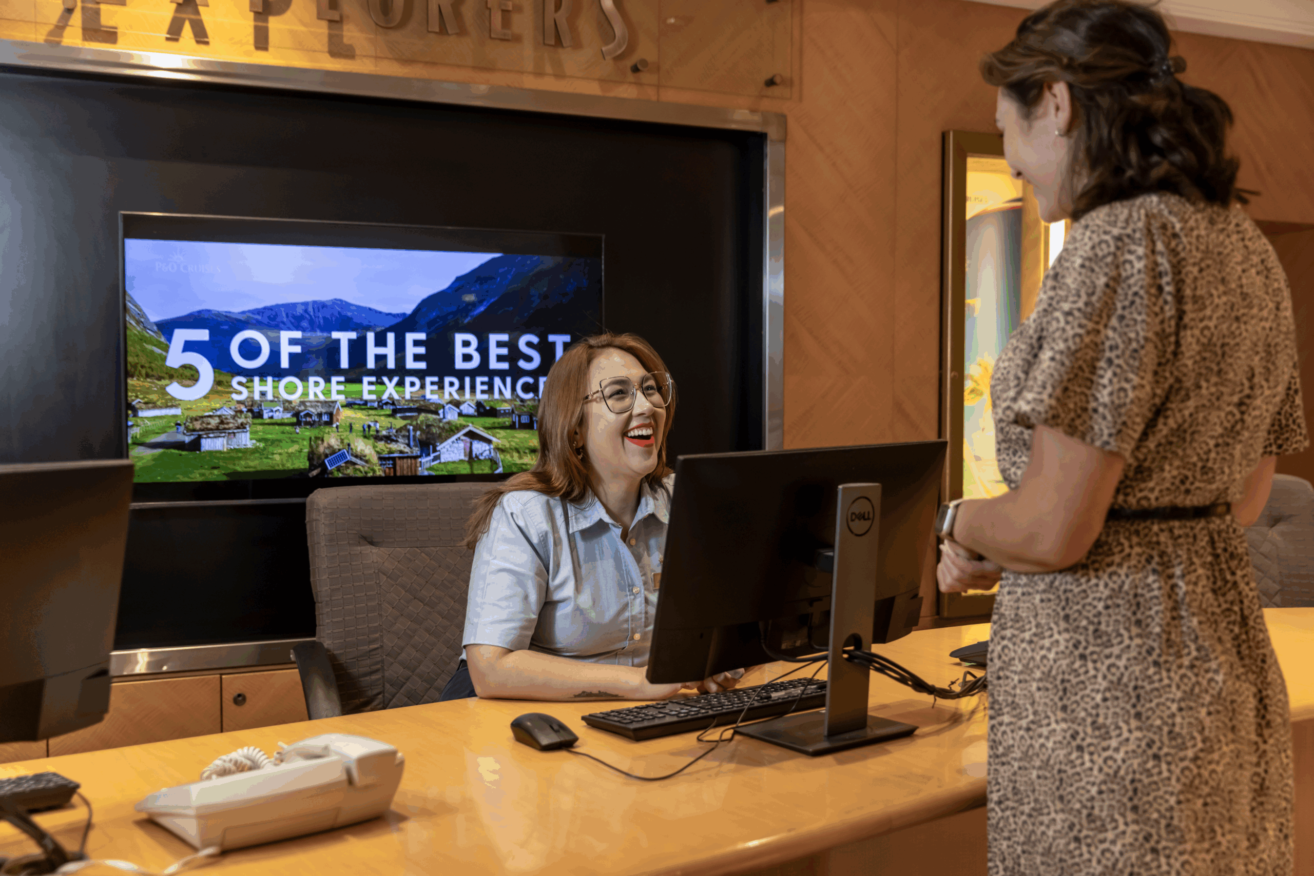 A receptionist smiles and talks to a guest at a reception desk. Behind them, a screen displays an image of mountains and cabins with the text 5 of the Best Shore Experiences. The setting appears welcoming and professional.