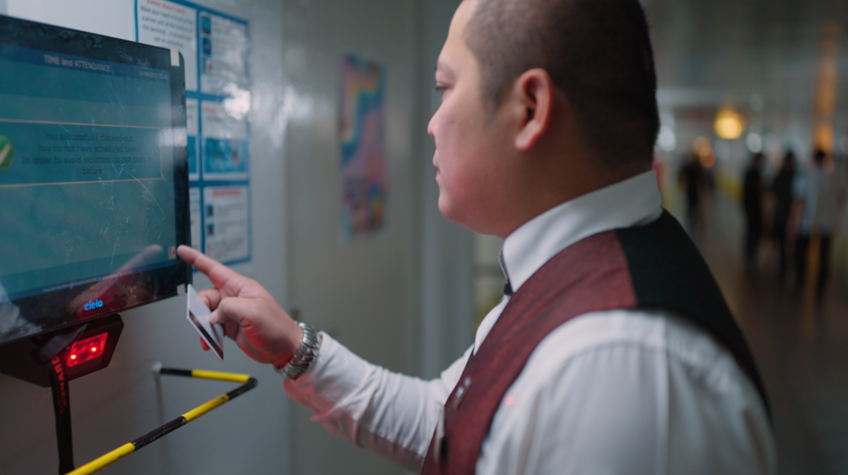 A man in a formal waistcoat and white shirt uses a touchscreen device mounted on a wall, holding a card in one hand. He appears to be in a corridor with information posters visible on the wall.