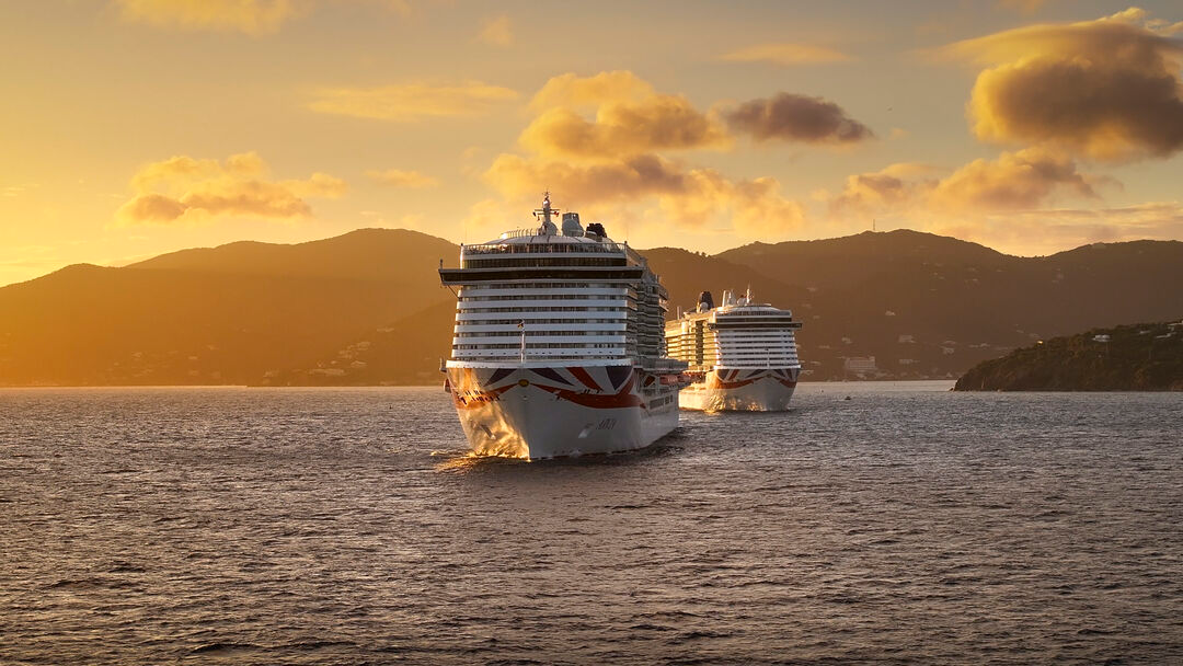 Two large cruise ships sail on calm ocean waters at sunset, with mountains silhouetted in the background and golden clouds scattered across the sky.