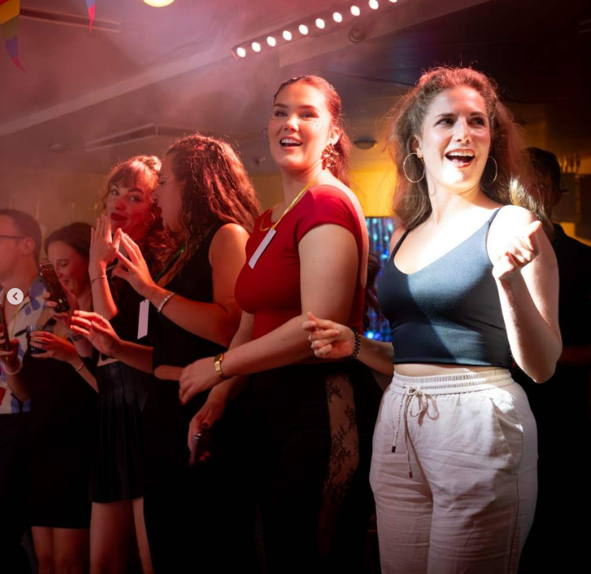 A group of young people stand closely together in a lively, colourful indoor setting, illuminated by bright lights. Two women in front are smiling and appear to be enjoying themselves, whilst others interact in the background.