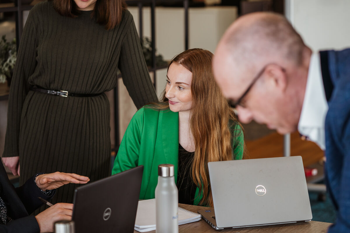 Four people have a discussion round a table with laptops and a notebook. A woman in a green blazer sits and smiles at a colleague whilst another person stands beside her and a man in the foreground leans forward.