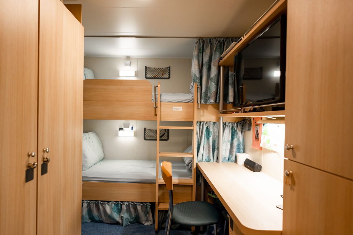 A tidy room with wooden furniture, featuring bunk beds, a desk with a chair, a TV mounted above the desk, lockers on the left, and patterned curtains by the beds.
