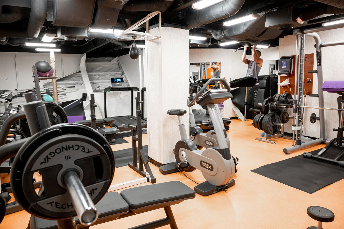 A gym with various fitness equipment, including a bench press, exercise bike, weights, and pull-up bar; a person is doing pull-ups in the background. The room has bright lighting and orange flooring.