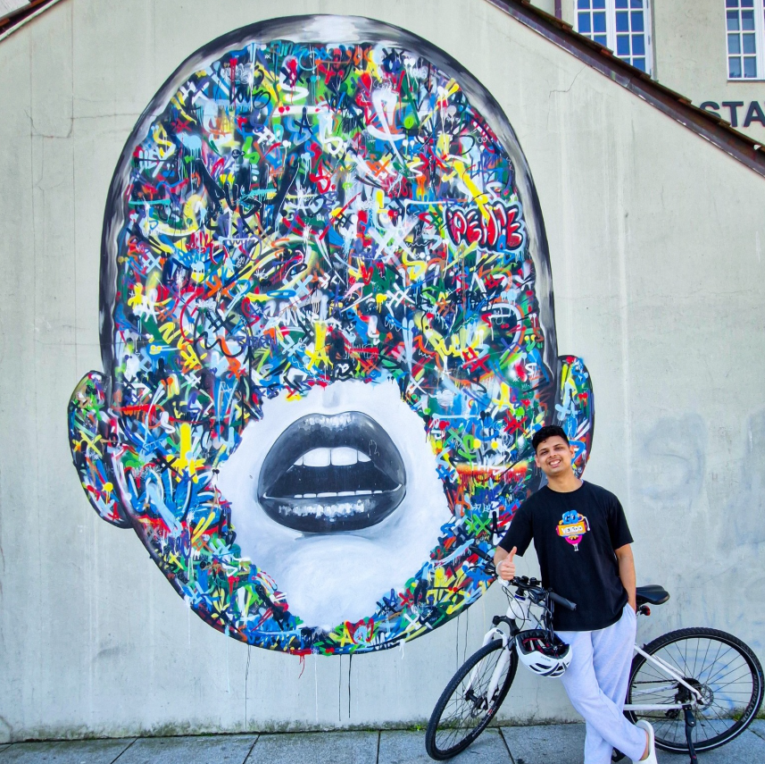A smiling person with a bicycle stands next to a mural of a large face covered in colourful, abstract graffiti lines on a grey wall. The mural's lips and ears are defined, but the rest of the face is obscured by paint.
