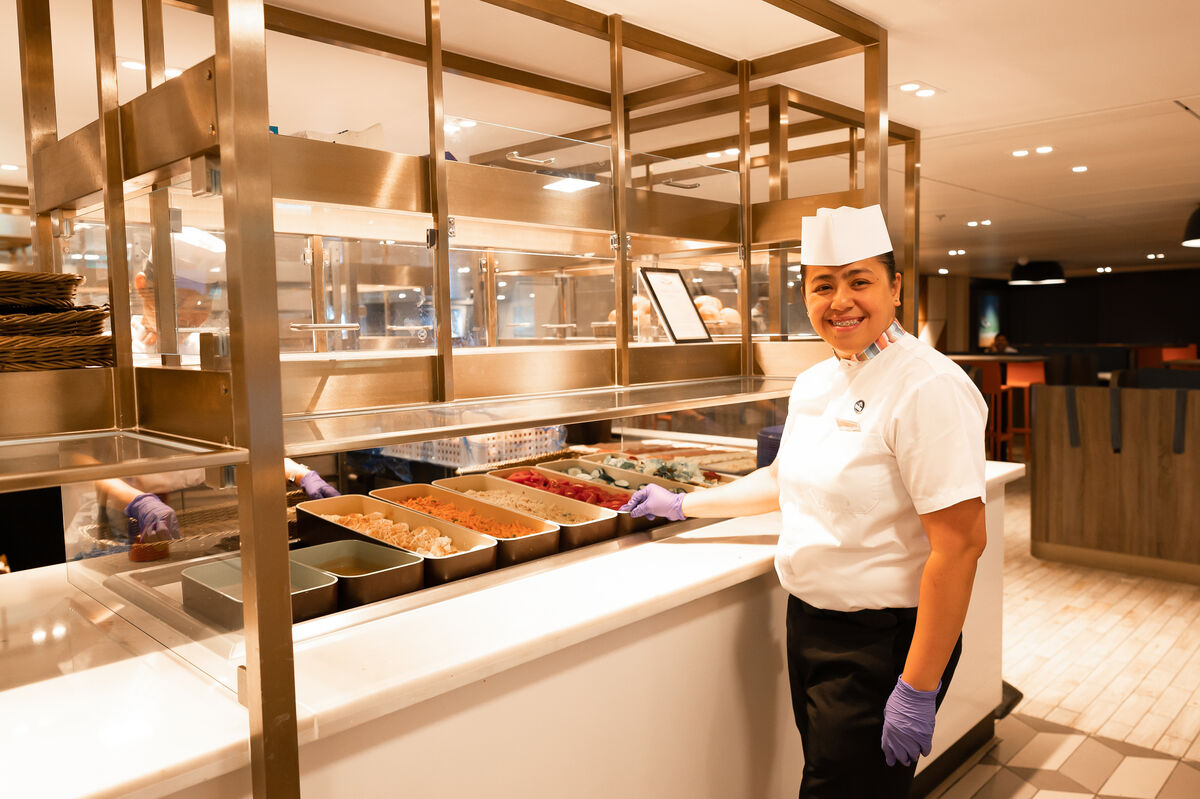 A smiling catering worker in uniform and gloves stands behind a counter with trays of various ingredients in a modern canteen or dining area.