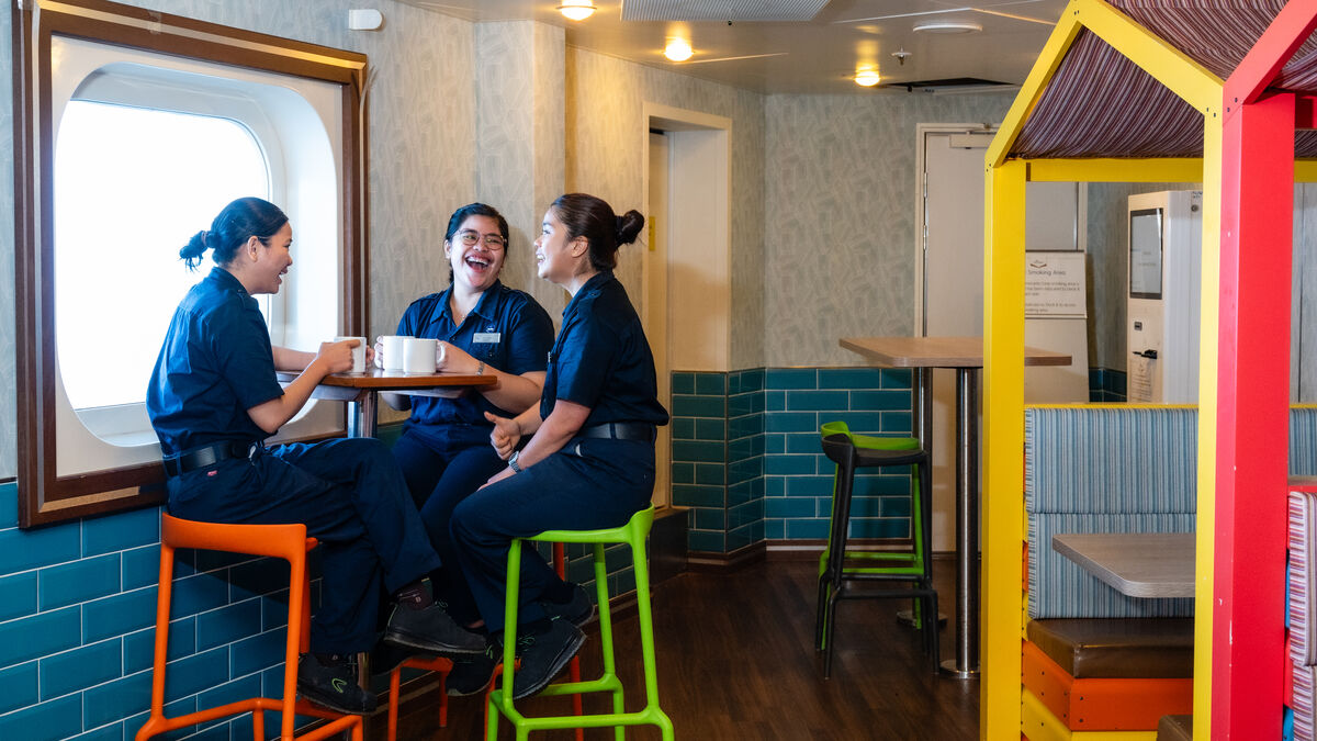 Three women in navy uniforms sit on colourful stools around a small table, smiling and chatting near a window in a bright, modern room with blue tile walls and playful décor.
