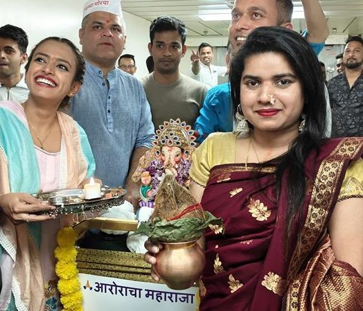 A group of people in festive attire celebrate Ganesh Chaturthi indoors. A woman in a maroon saree holds a kalash, whilst another woman holds an aarti plate. A Ganesha idol decorated with flowers is in the background.