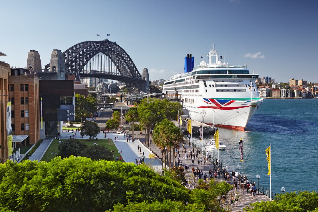 A large cruise ship docked at Circular Quay in Sydney, Australia, with the Sydney Harbour Bridge in the background and people walking along the waterfront promenade.