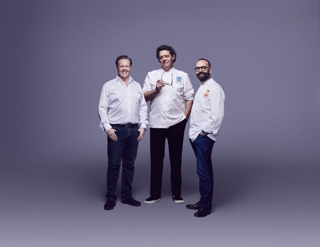 Three men stand together against a plain purple background, all smiling. Two wear white chef jackets and dark trousers, while the man on the left wears a white shirt and dark jeans.
