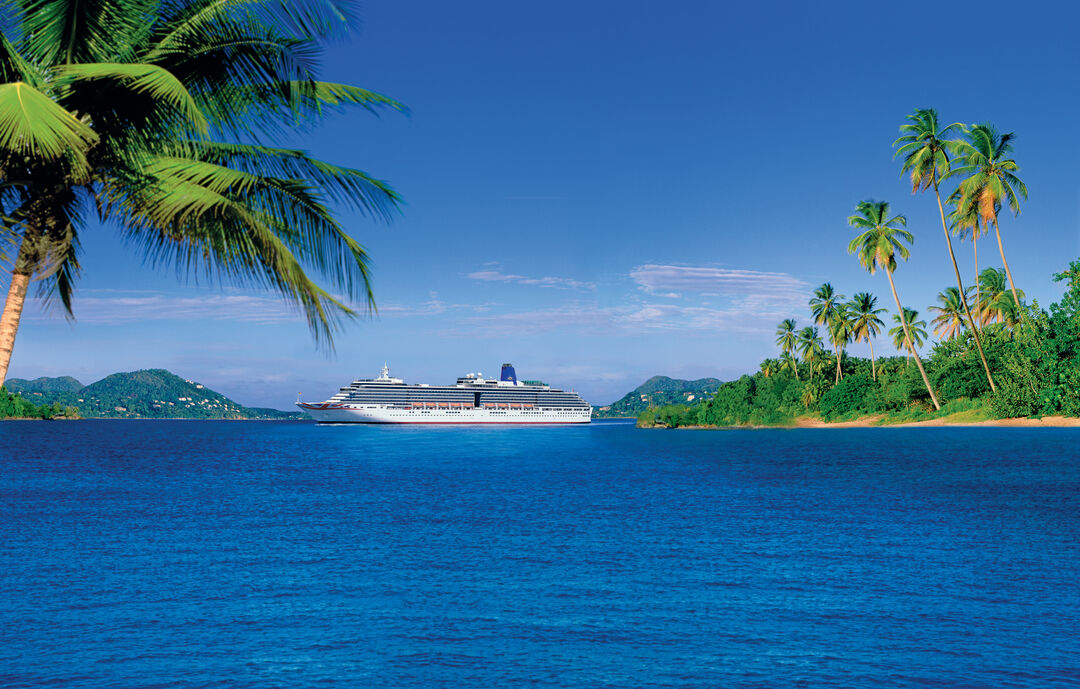 A large cruise ship sails on bright blue tropical waters, surrounded by lush green palm trees and distant islands under a clear blue sky.