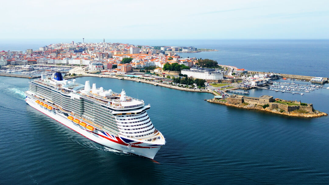 A large white cruise ship sails into a coastal city harbour with historic buildings, a marina, and a fortified structure visible on the shoreline under a clear sky.