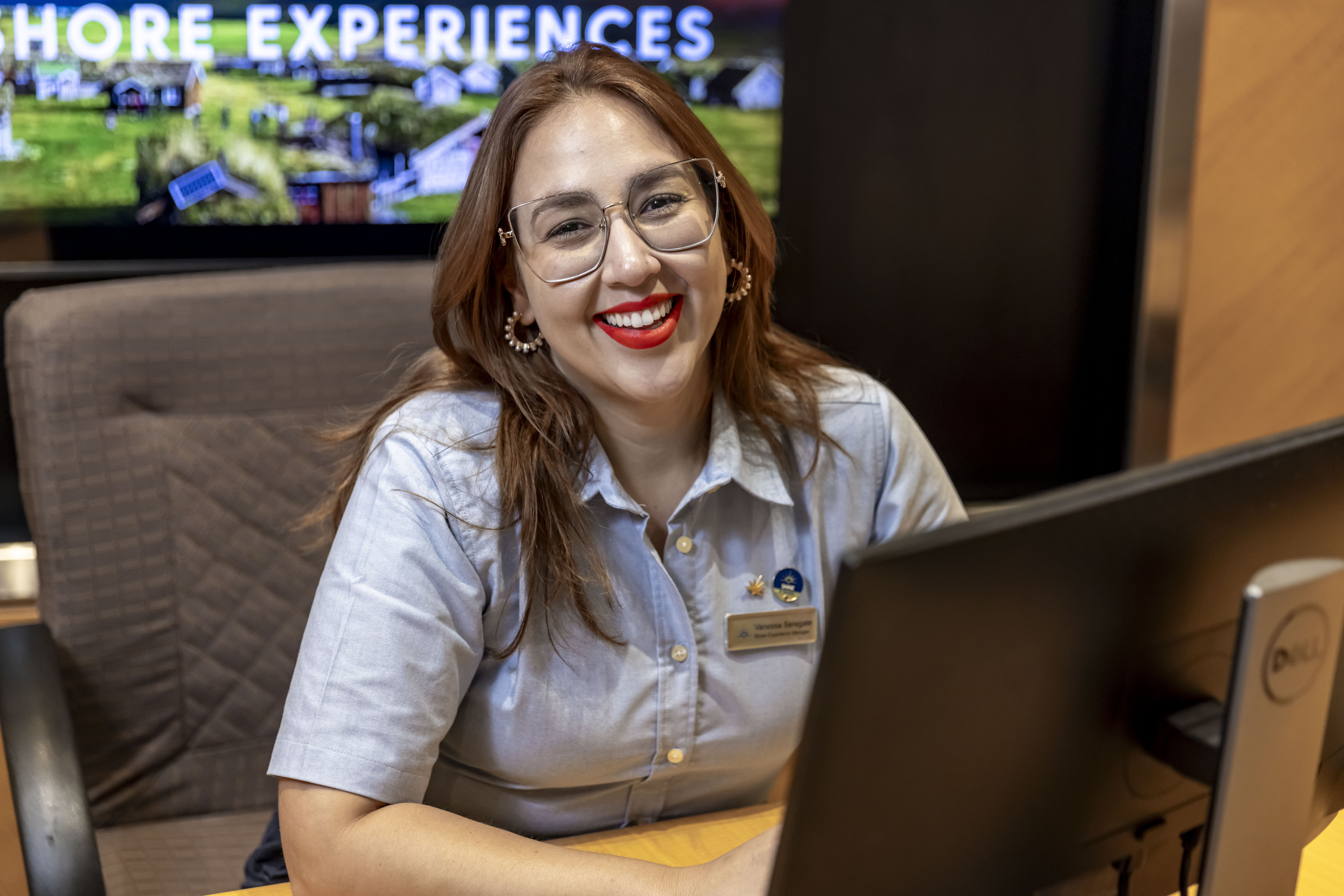 A smiling woman with glasses and red lipstick sits at a desk in front of a computer, wearing a light blue shirt and a name badge. A sign behind her reads SHORE EXPERIENCES.
