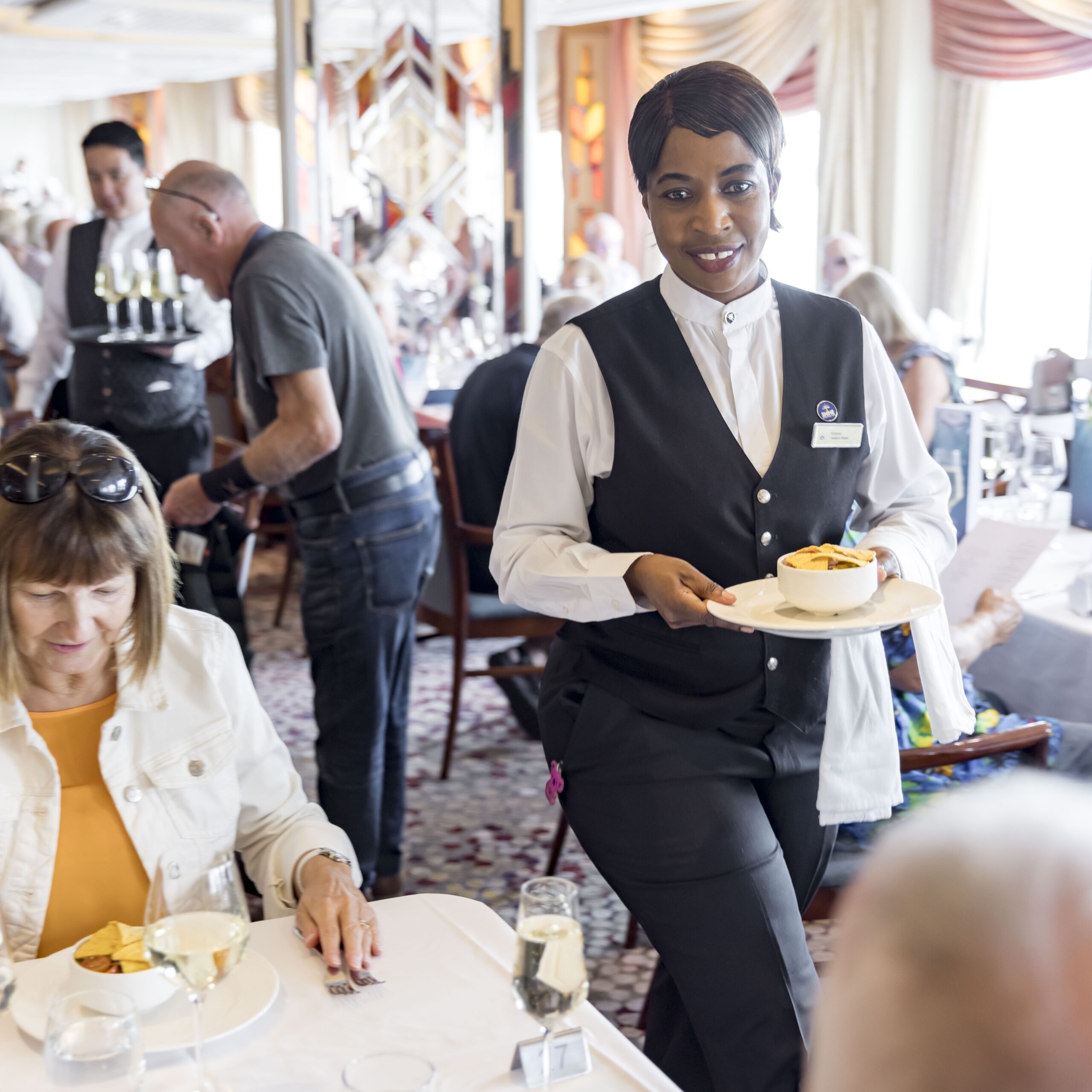 A smiling waiter in a black waistcoat and white shirt carries a plated pudding to a table in a busy, elegant restaurant, where guests are seated and dining.