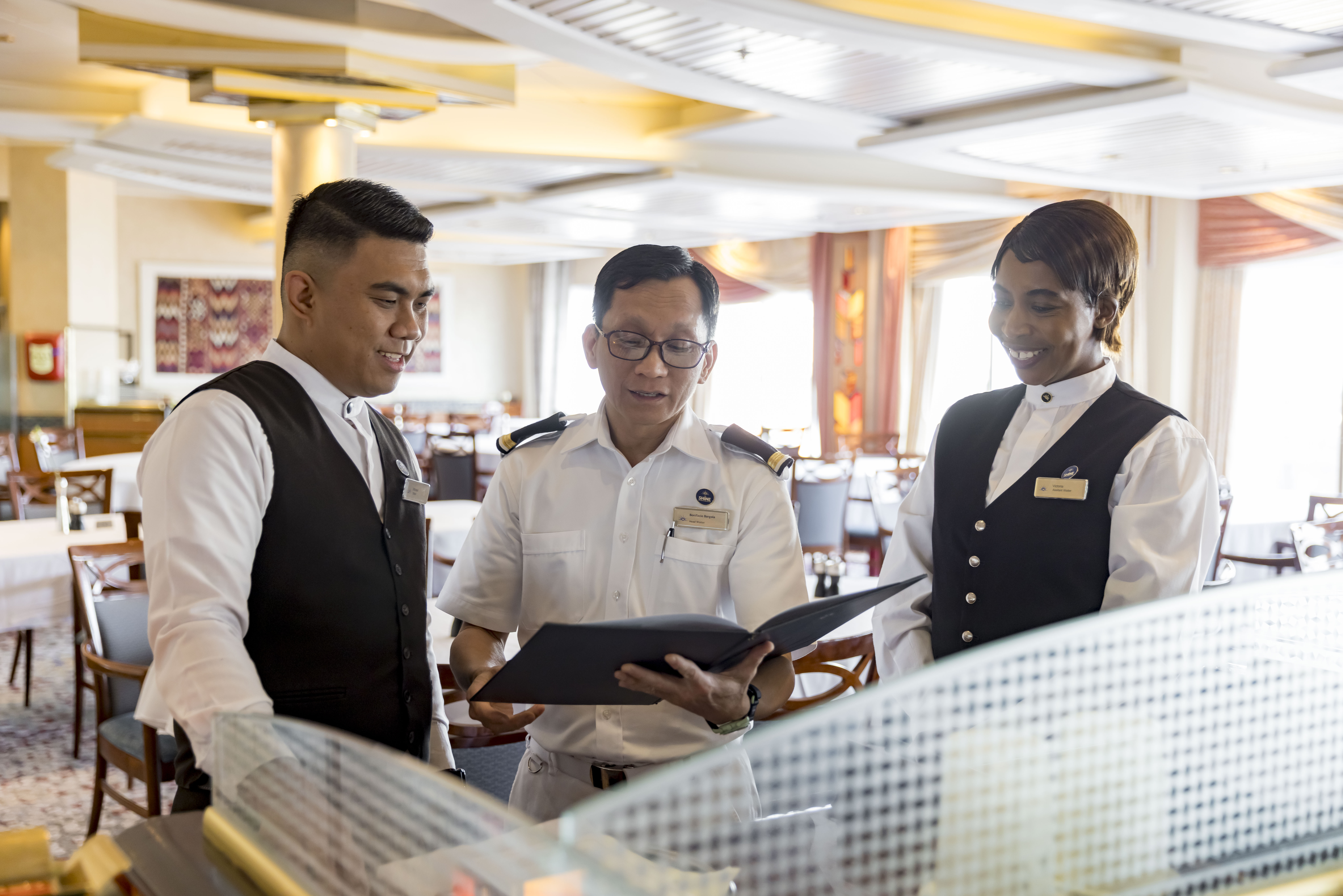 Three restaurant staff members in uniform stand together, smiling and looking at a folder. They appear to be discussing something inside a bright, elegant dining area with neatly arranged tables and chairs.