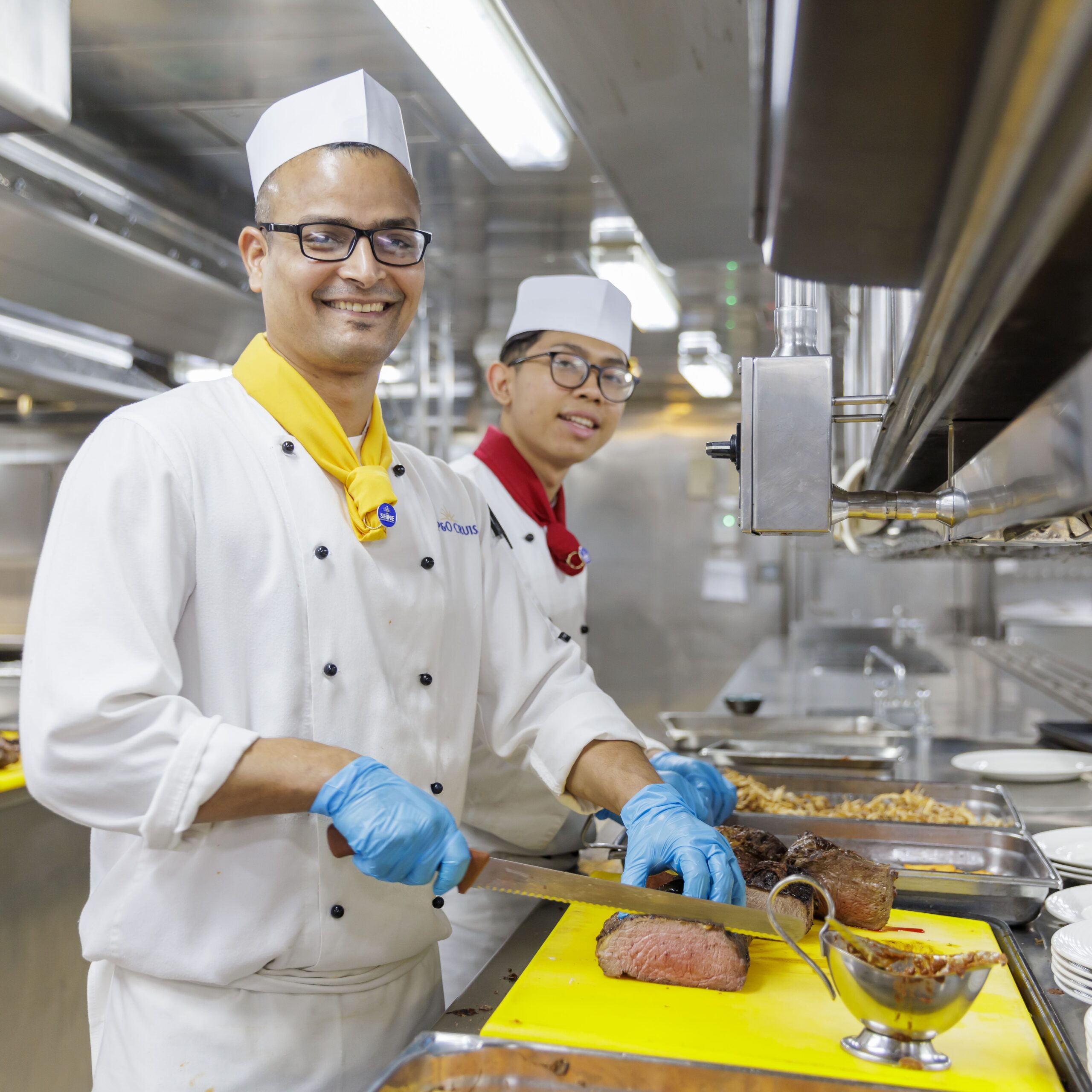 Two chefs in white uniforms and hats slice cooked meat on a yellow chopping board in a professional kitchen. One chef smiles at the camera whilst the other works in the background.