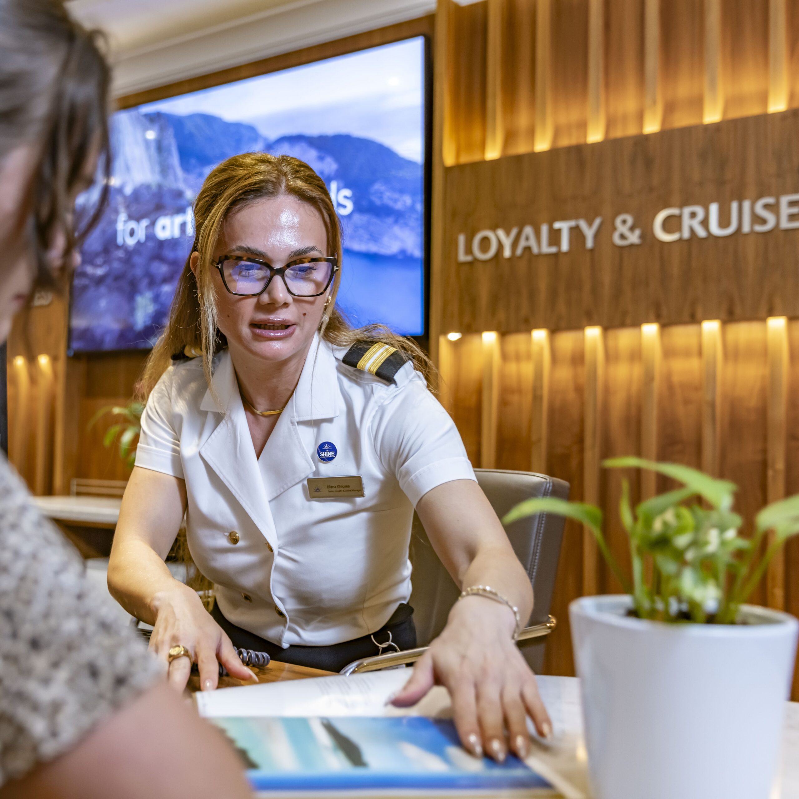 A woman in a cruise officer uniform sits at a desk, talking to a guest. She is gesturing towards a brochure. Behind her, a wooden wall sign reads LOYALTY & CRUISE. There are plants and a screen in the background.