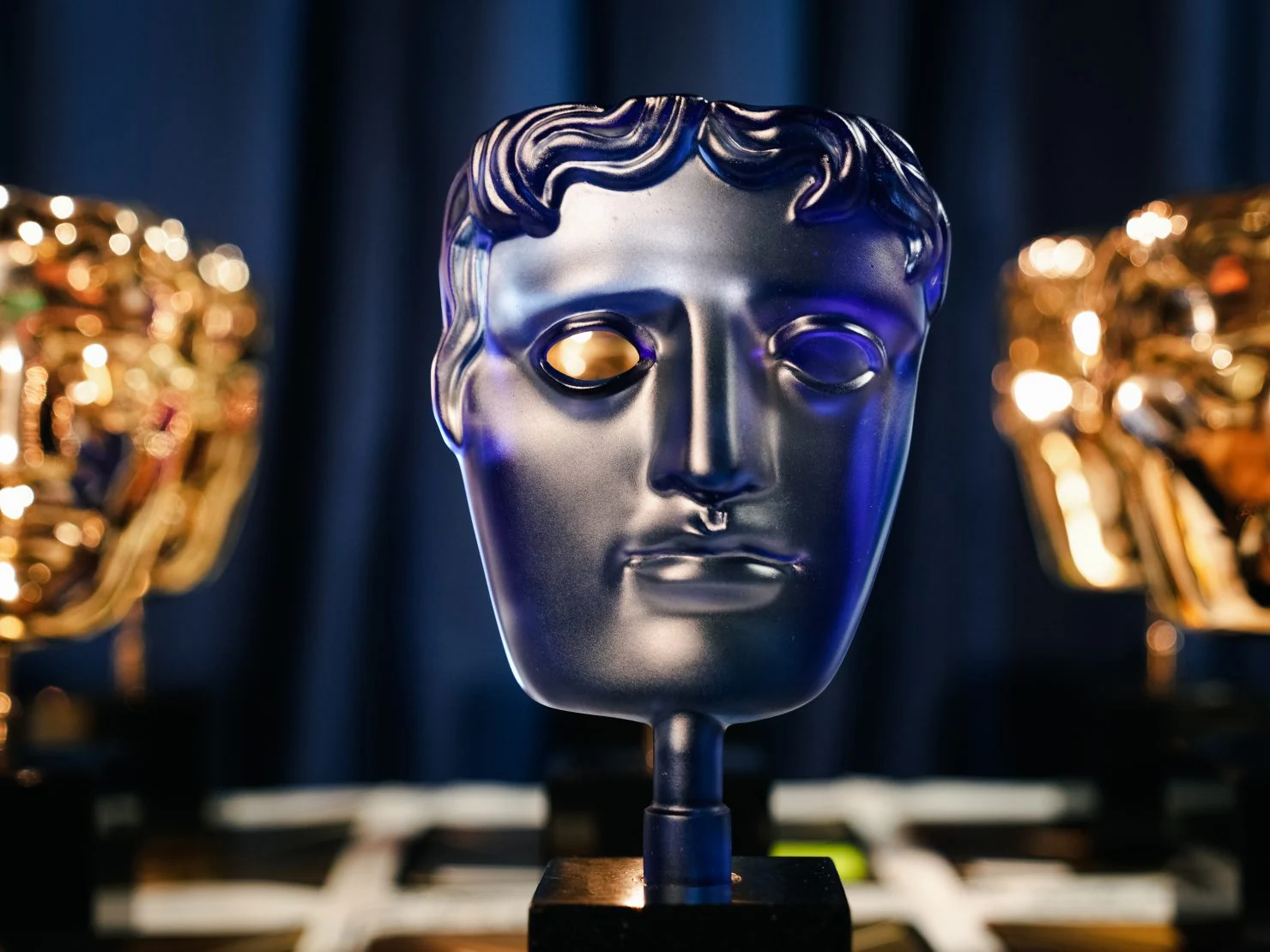 A close-up of a blue-tinted BAFTA mask trophy in focus, with two gold BAFTA trophies blurred in the background, set against a dark backdrop.