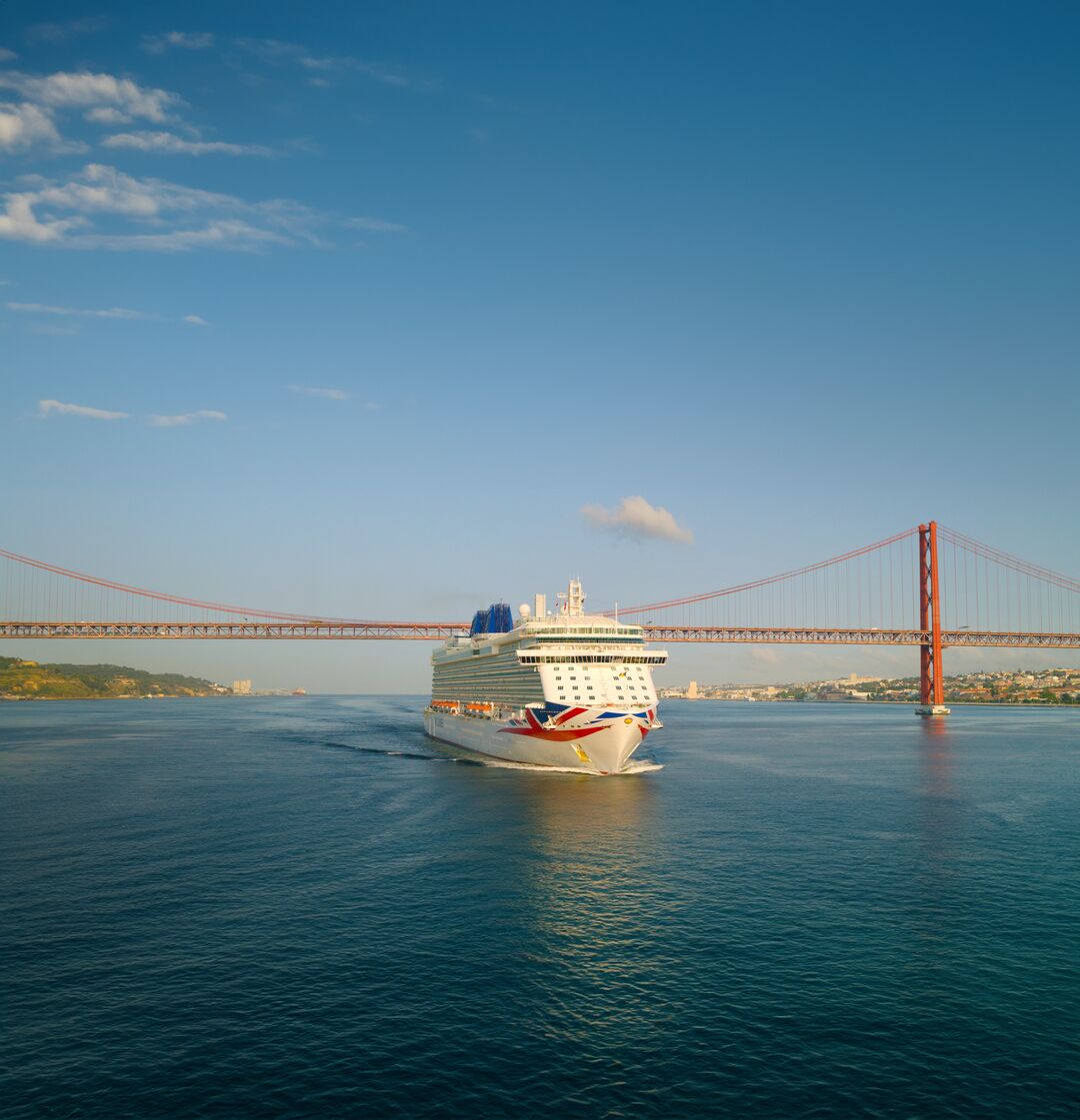 A large cruise ship sails under a red suspension bridge on calm blue water, with a clear sky and cityscape in the background.