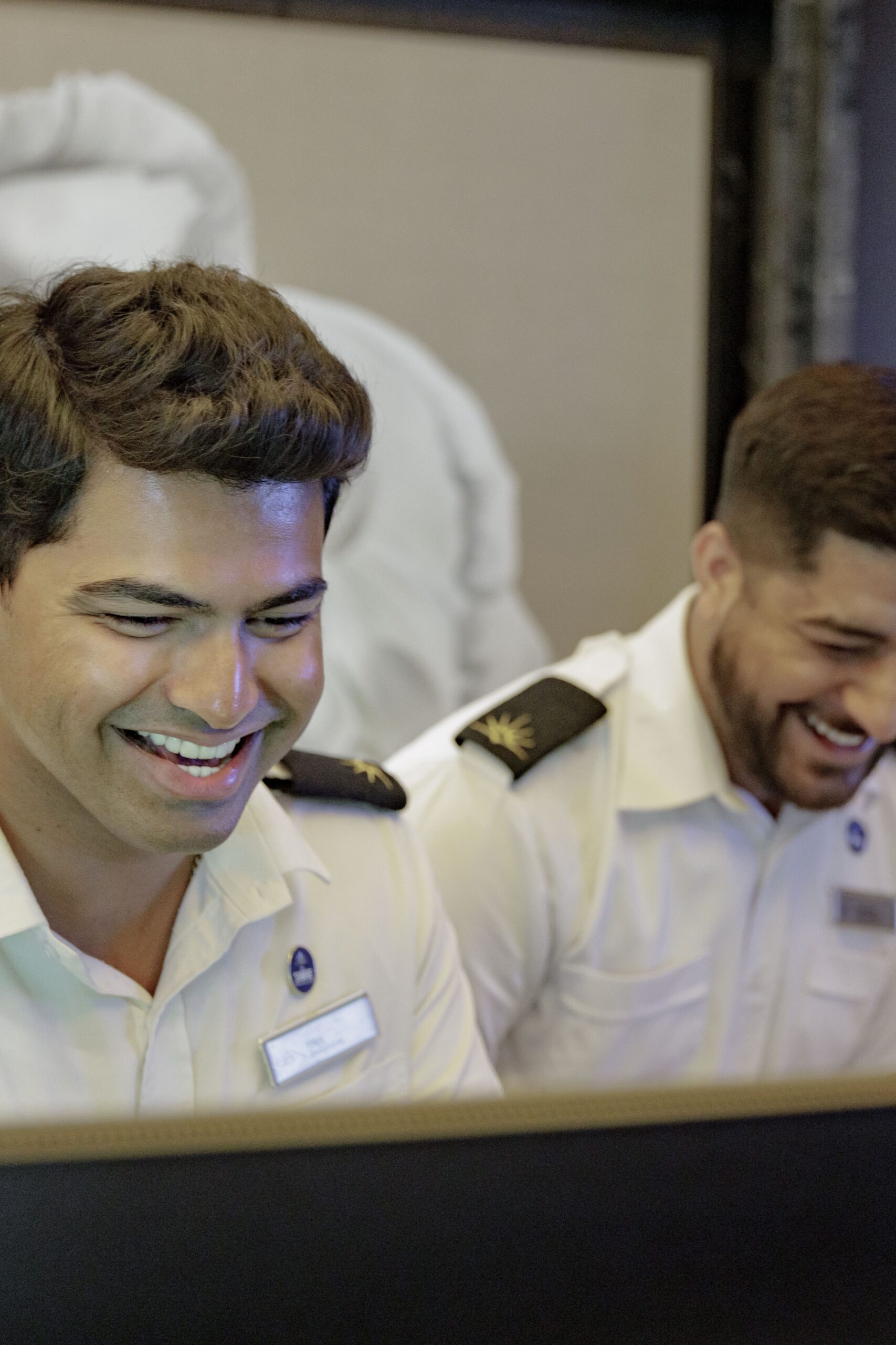 Two men in white uniforms with shoulder epaulettes are smiling and looking at a computer screen. They appear to be enjoying their work and are in a professional indoor setting.