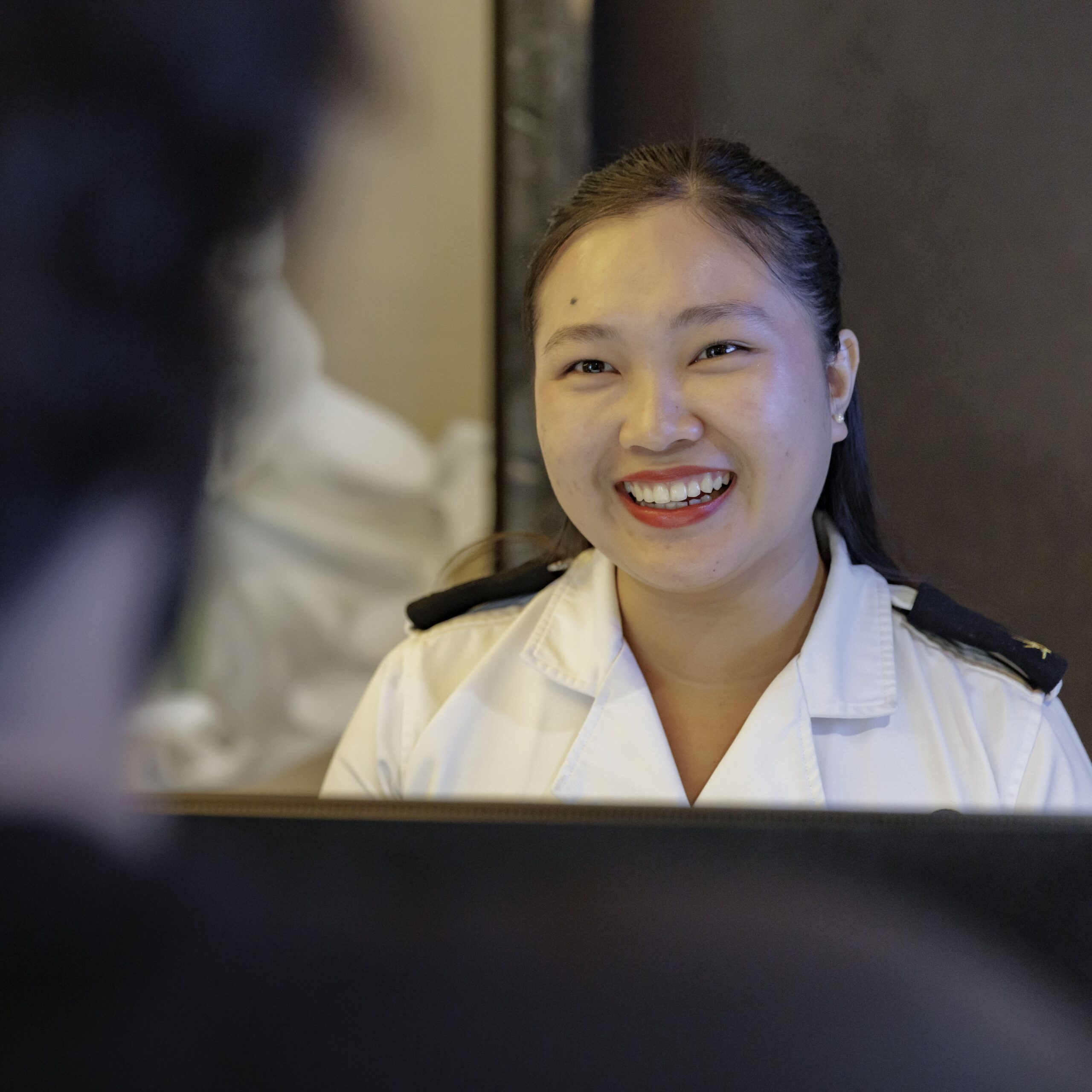 A woman in a white uniform is smiling warmly whilst speaking to a person across a counter, suggesting a friendly customer service interaction.