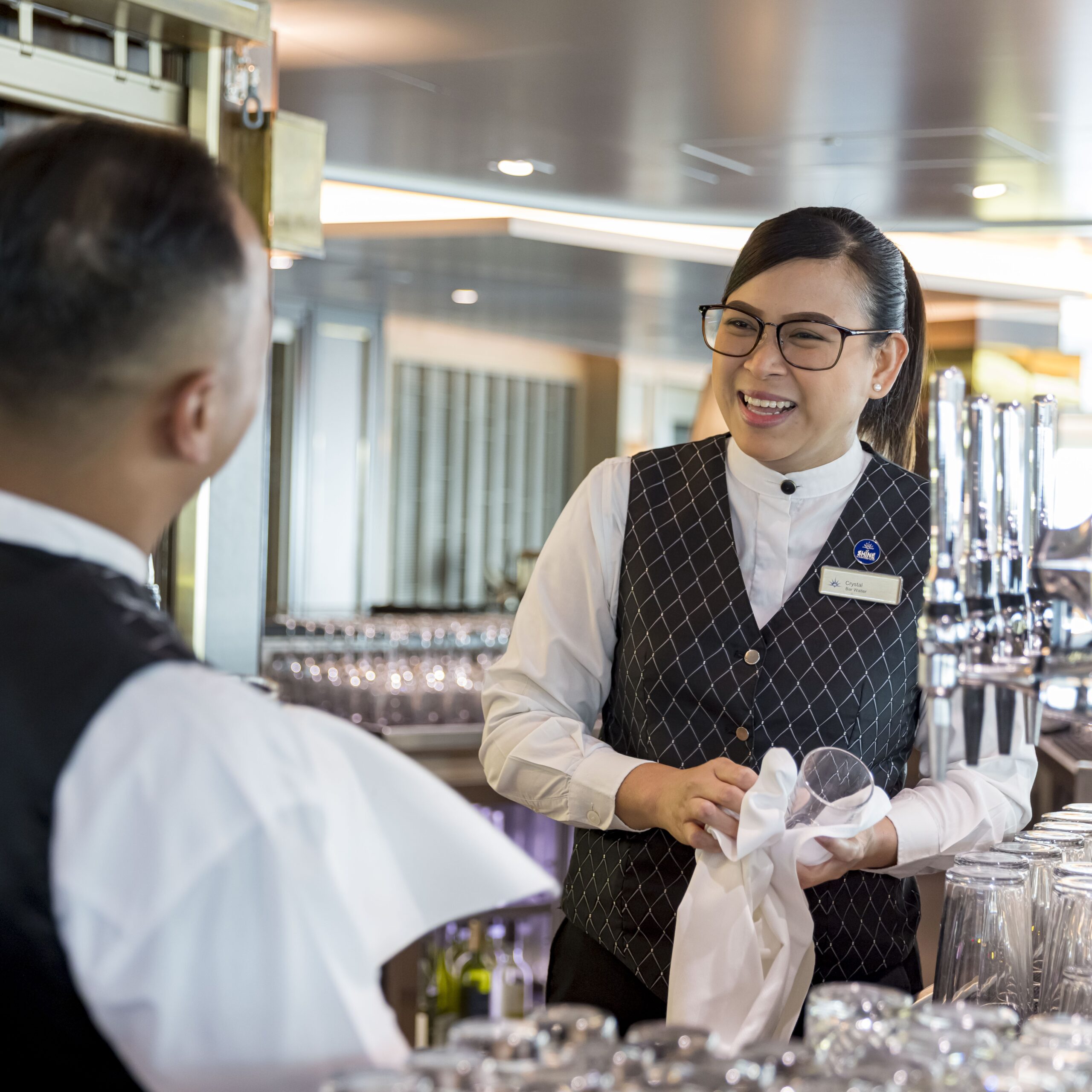 Two bartenders in uniform stand behind a bar, smiling and conversing. One person dries a glass with a tea towel, while the background shows shelves filled with glassware and drink dispensers.