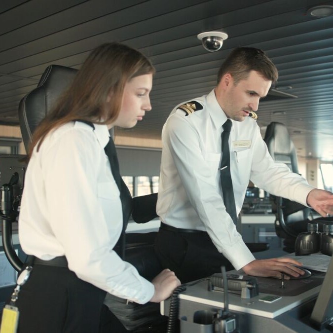 Two people in white uniforms stand at a control panel on a ship’s bridge. One is seated and observing, whilst the other is standing and interacting with touchscreen controls. Large windows surround them.
