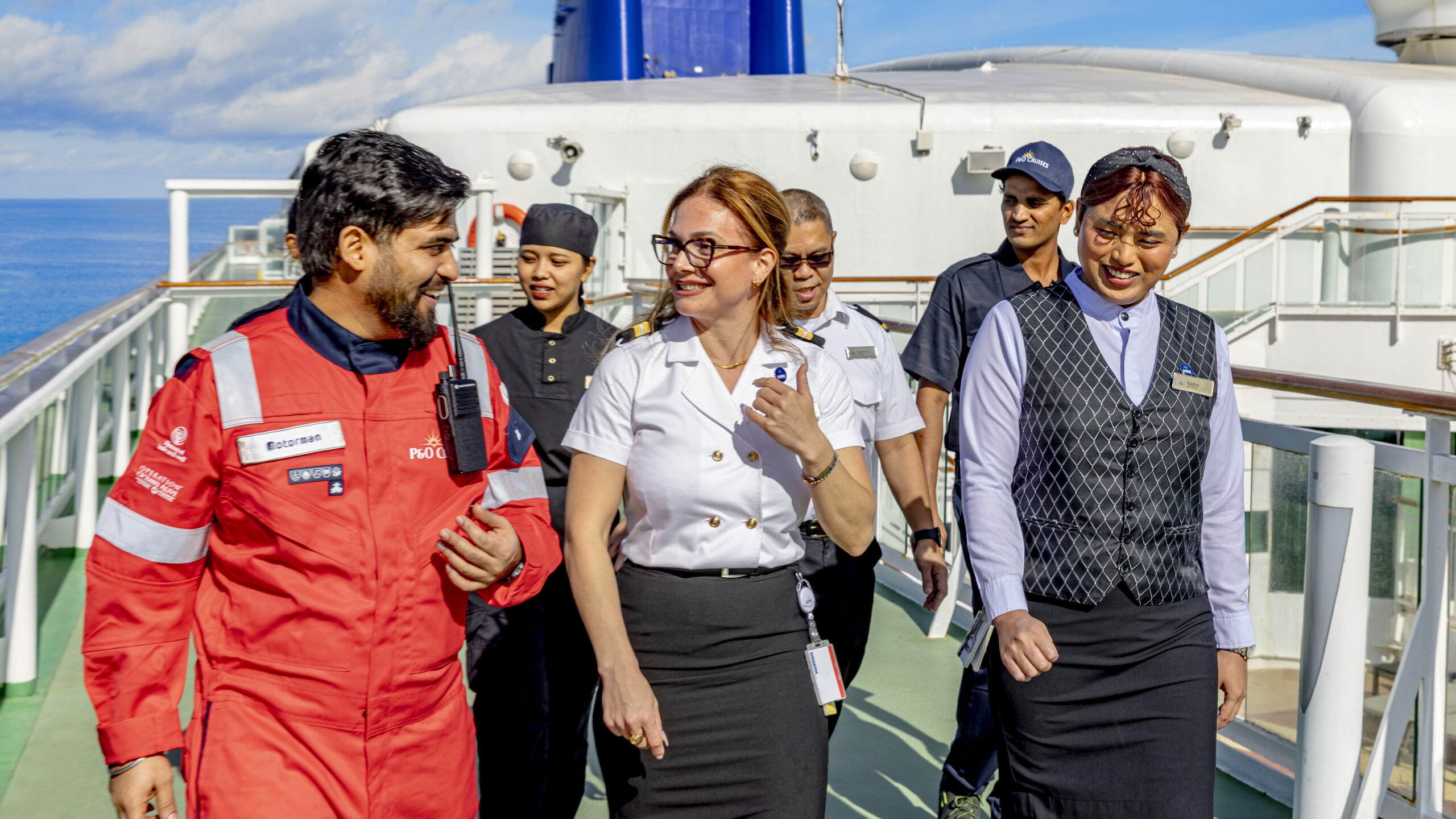 A group of cruise ship crew members in uniforms walk and talk together on the deck, smiling and enjoying a sunny day. The ship’s railings and exterior structure are visible in the background.