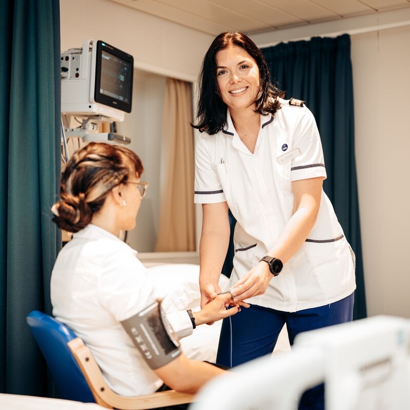 A nurse in white uniform smiles whilst checking the blood pressure of a seated patient in a medical room, using a monitor and cuff. The patient faces away from the camera.