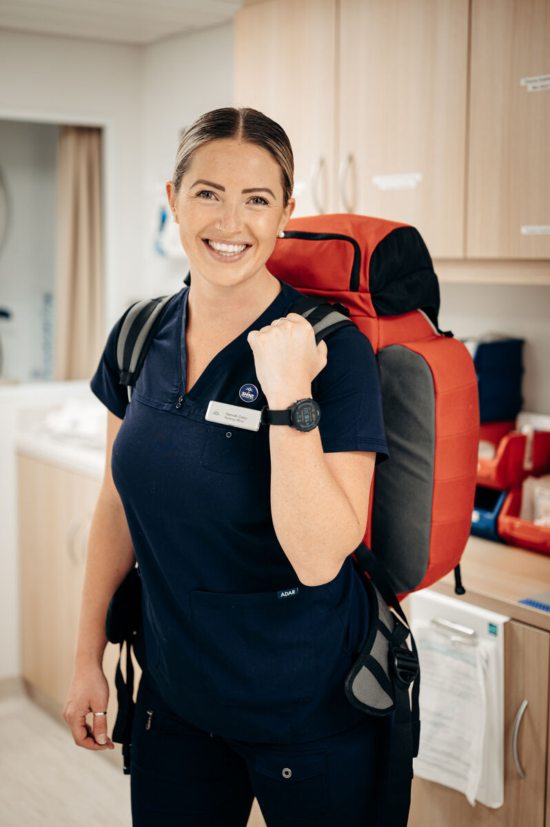 A smiling healthcare worker in navy scrubs stands indoors, wearing a name badge and a large red rucksack, with medical supplies visible in the background.