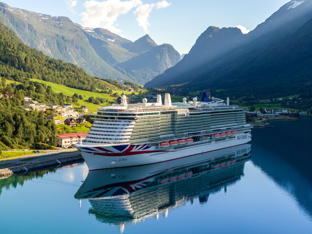 A large cruise ship sails on a calm fjord, reflecting in the water, with green hills, small buildings, and tall mountains in the background under a partly cloudy sky.