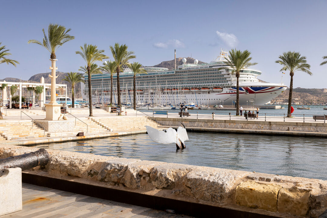 A large cruise ship is docked near a sunny waterfront lined with palm trees and stone steps. In the foreground, there is a small canal with a white swan sculpture and an old cannon on a stone wall.