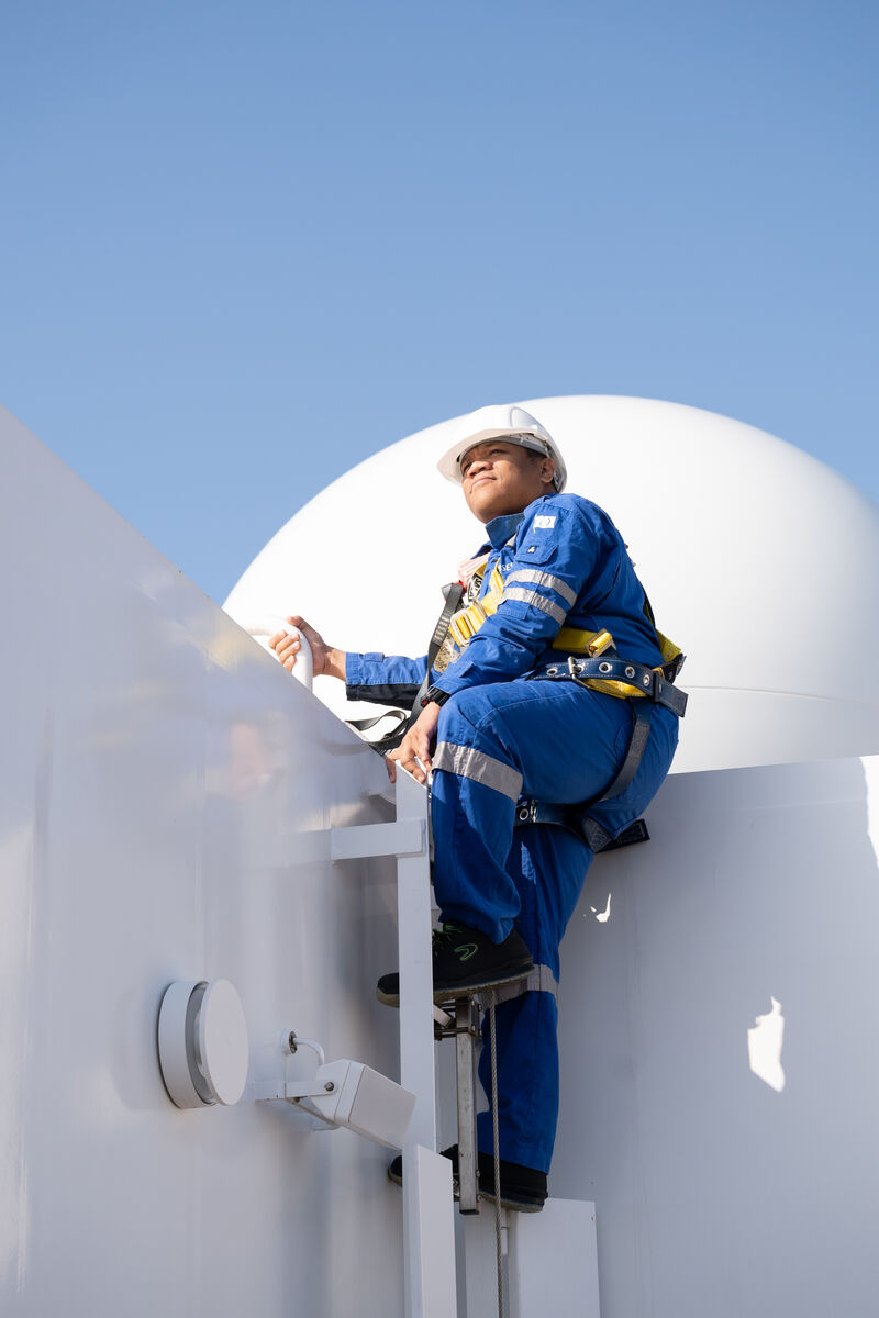 A person in blue overalls, safety harness, and a white hard hat climbs a metal ladder attached to a white structure under a clear blue sky.