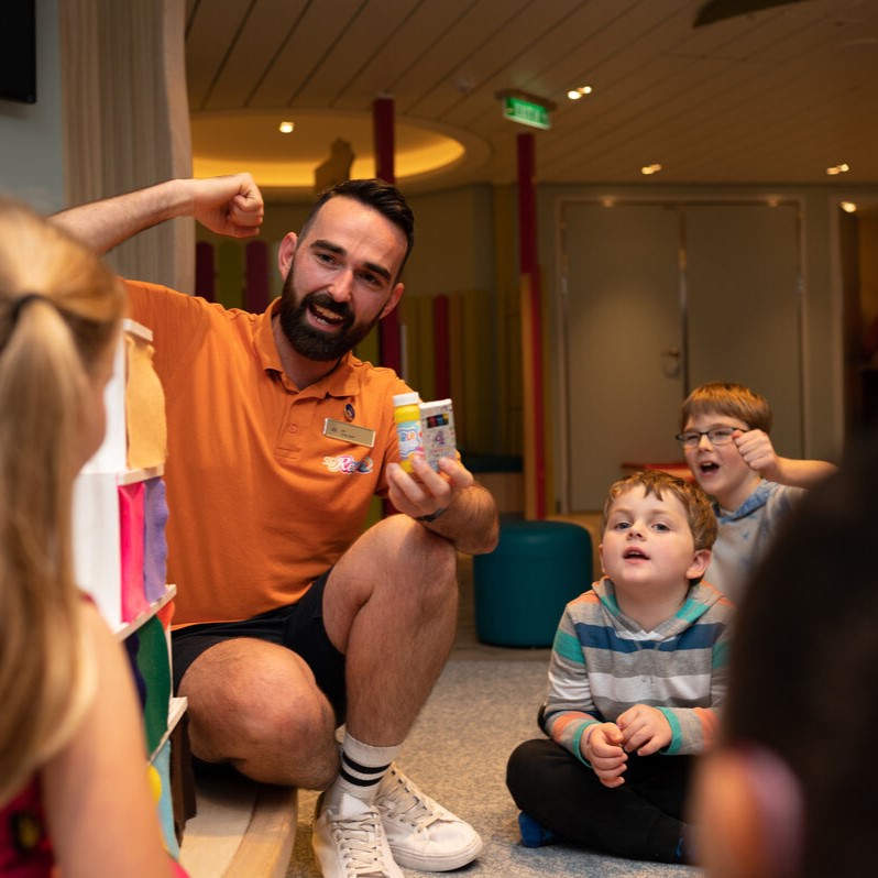 A man kneels and smiles enthusiastically whilst holding up two bottles, engaging a group of young children sitting on the floor in a colourful indoor setting.