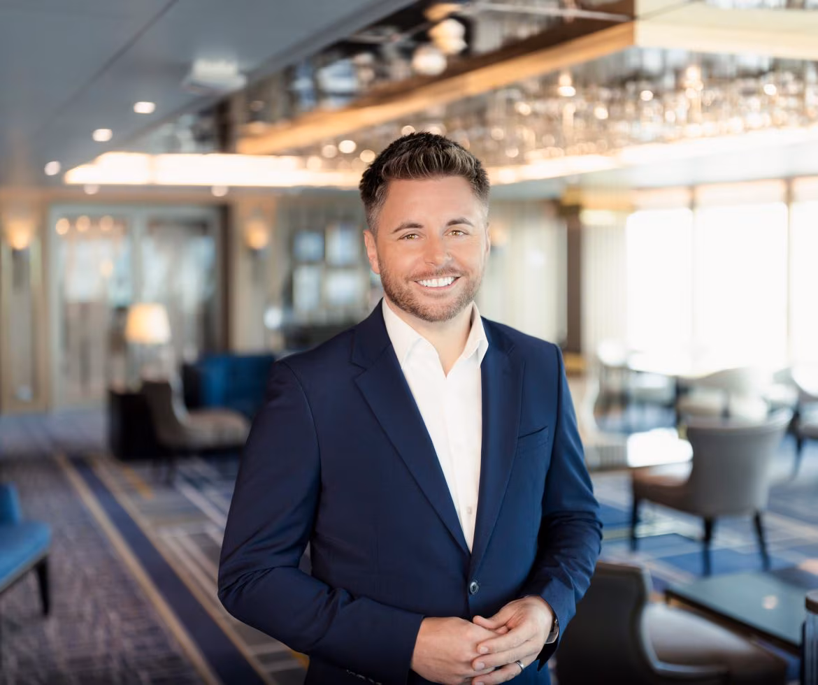A man in a navy blue suit and white shirt stands smiling in a modern, well-lit lounge with elegant decor, armchairs, and large windows in the background.