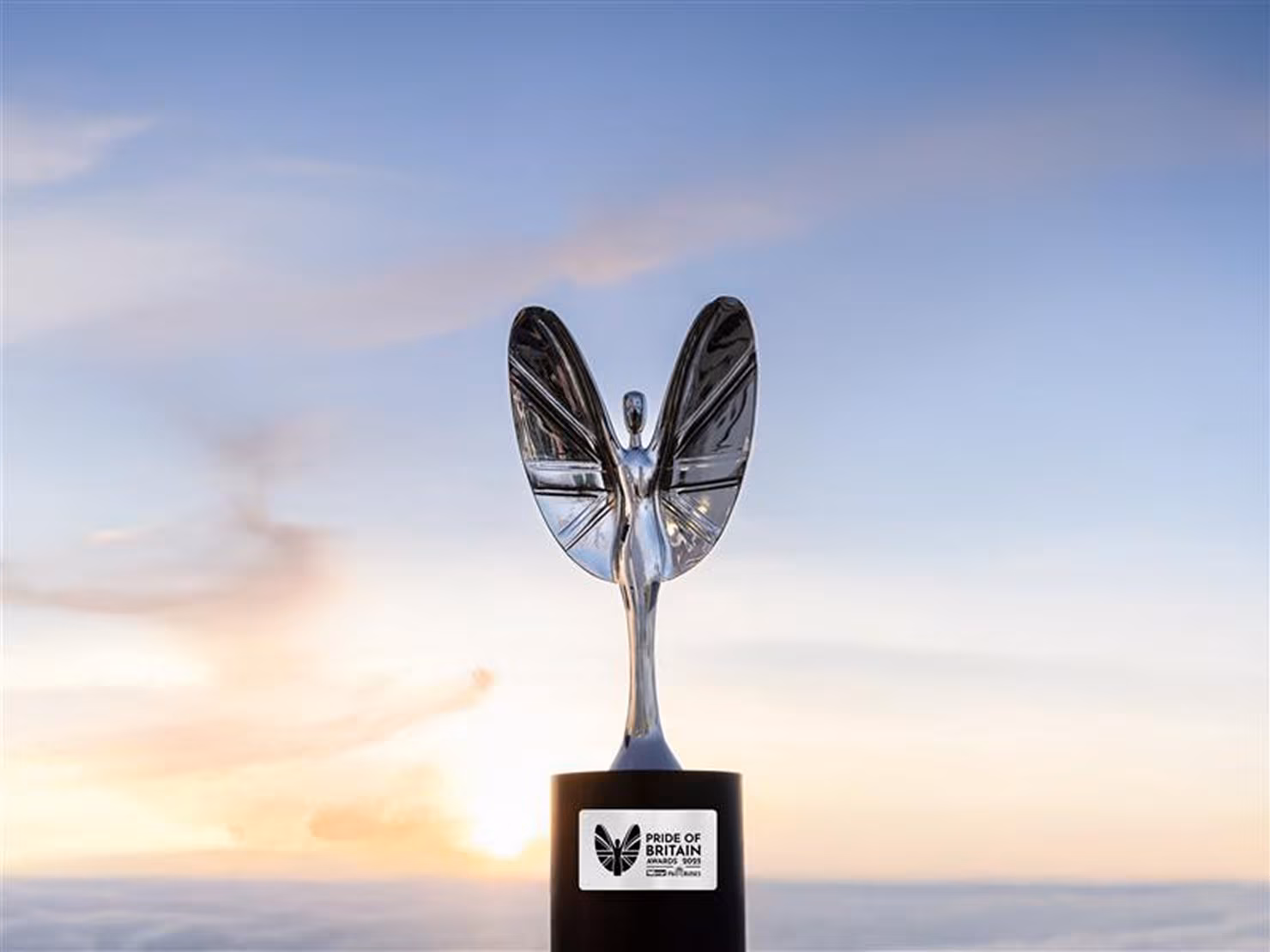 A silver angel-shaped Pride of Britain award trophy stands on a black base against a soft, pastel sky at sundown.