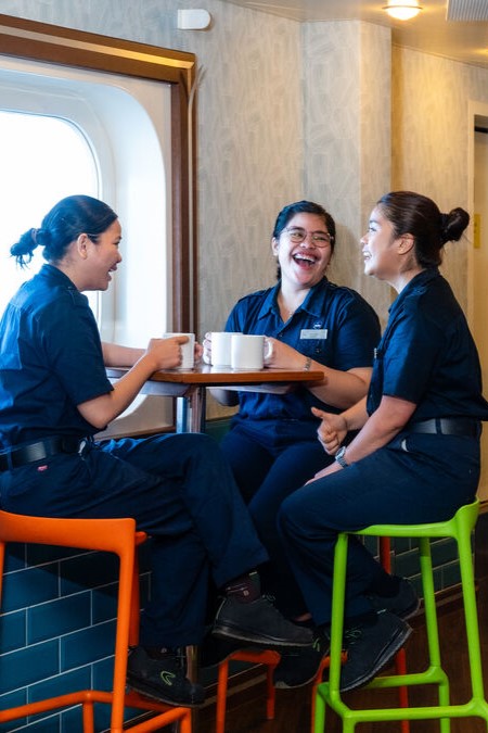 Three women in navy blue uniforms sit on colourful stools around a table, smiling and laughing together whilst holding mugs in a brightly lit room with a window.