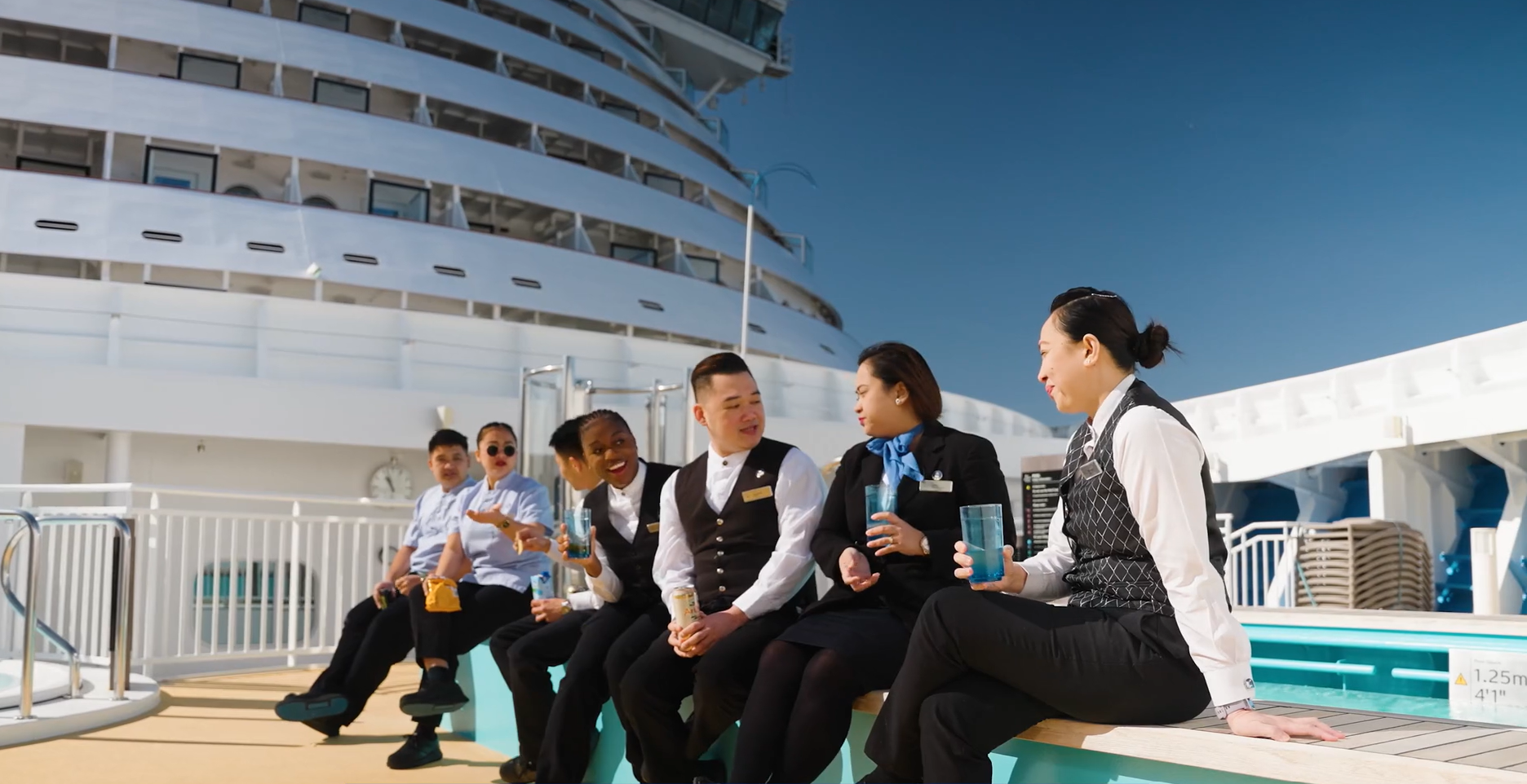 Six cruise ship staff members in uniform sit together on a bench by a swimming pool, smiling and talking during a break, with the cruise ship and clear blue sky in the background.