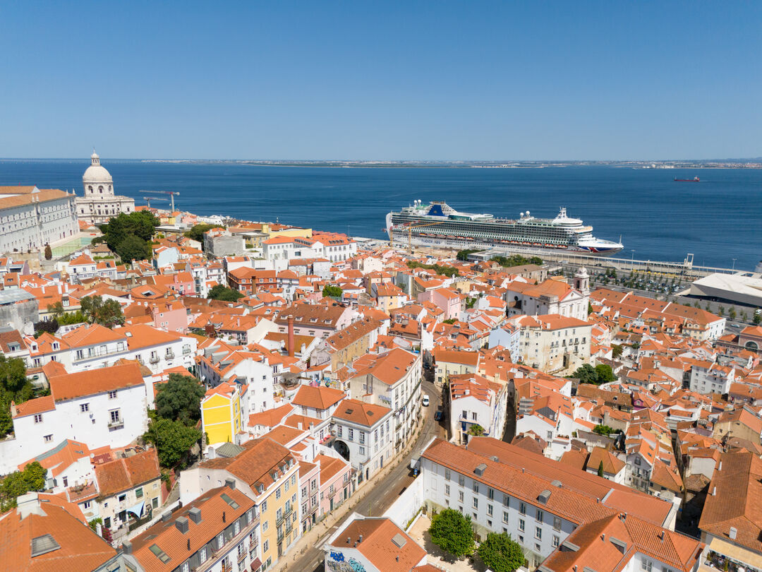 Aerial view of Lisbon’s Alfama district with red-roofed buildings, the National Pantheon, and a large cruise ship docked by the Tagus River under a clear blue sky.