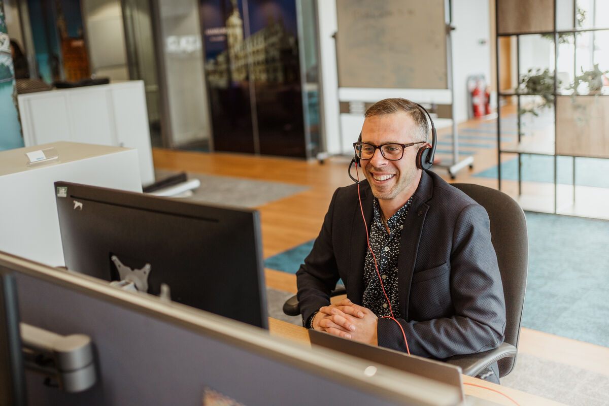 A man wearing glasses and a headset smiles whilst sitting at a desk in an office, looking at a computer monitor, appearing engaged in a video call or virtual meeting.