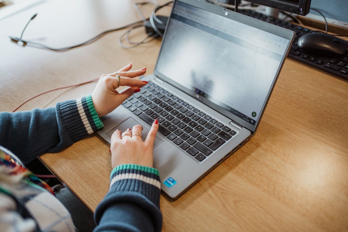 A person with rings on their fingers types on a laptop at a wooden desk, with another computer mouse and monitor visible in the background.