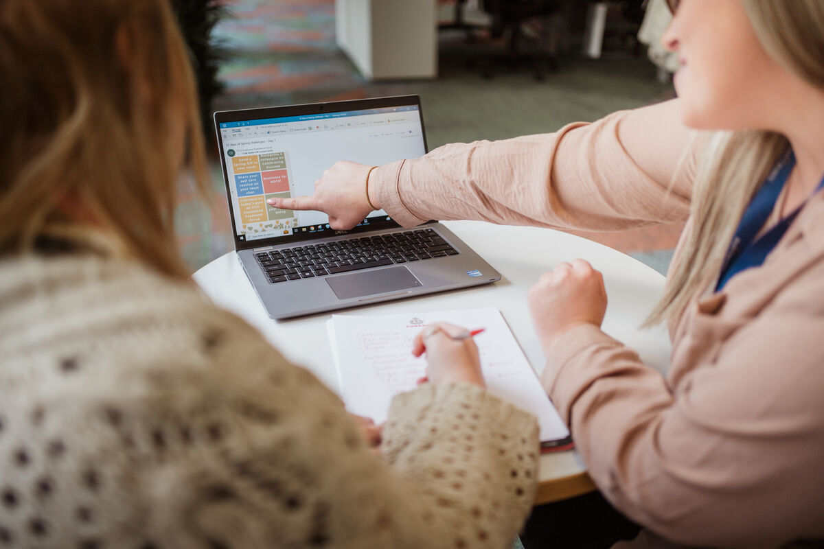Two women sit at a table with a laptop. One points at the screen displaying colourful notes, while the other writes in a notebook. They appear to be discussing or collaborating on work.